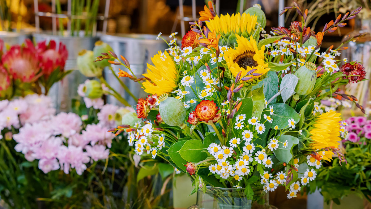 A vibrant summer flower bouquet featuring yellow sunflowers, white daisies, pink protea, and green foliage, arranged in a clear glass jar on a wooden table in a flower shop.